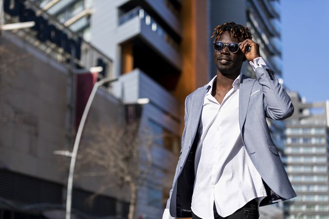 Black man in elegant suit & sunglasses walking in the street while adjusting his glasses on a sunny day