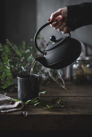 Woman pouring green tea in mug on wooden table with green herb