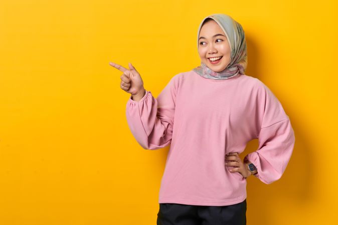 Smiling Muslim woman in headscarf pointing to space beside her with hand on hip