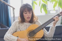 Female singing and strumming guitar in living room of bright loft with plants in background 0yEP70
