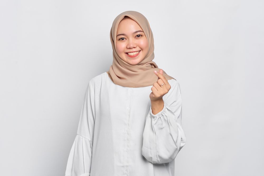 Muslim woman in headscarf and light blouse and smiling and making heart shape with fingers