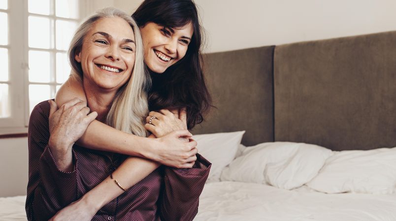 Smiling adult woman holding her mother from behind sitting on bed