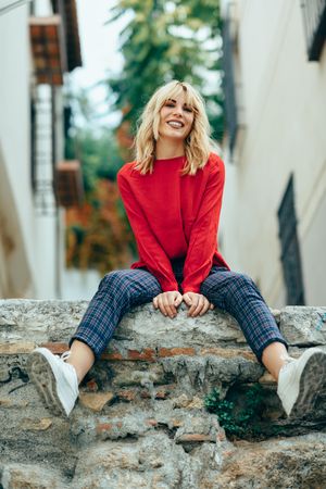 Happy blond woman sitting on stone wall with her legs dangling