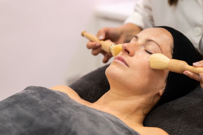 Crop beautician massage with brushes on female's cheeks in beauty salon