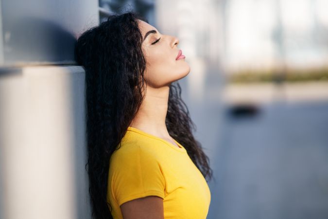 Profile of female with curly hair leaning back with eyes closed on grey wall outside, copy space