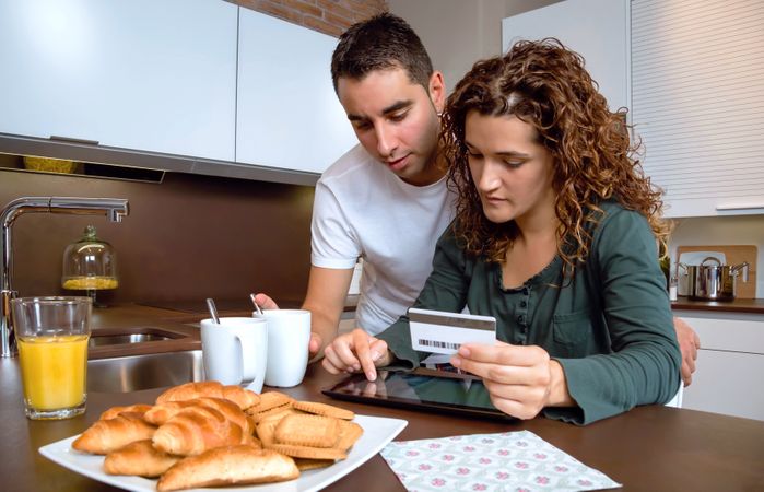 Couple having breakfast in kitchen while paying for something with tablet