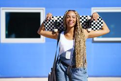 Joyful female in denim overalls standing with skateboard behind head in front of blue building
