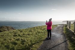 Woman standing on country road and taking picture of sea with smartphone