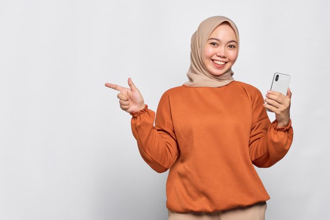 Smiling Muslim woman holding smart phone and pointing to her side