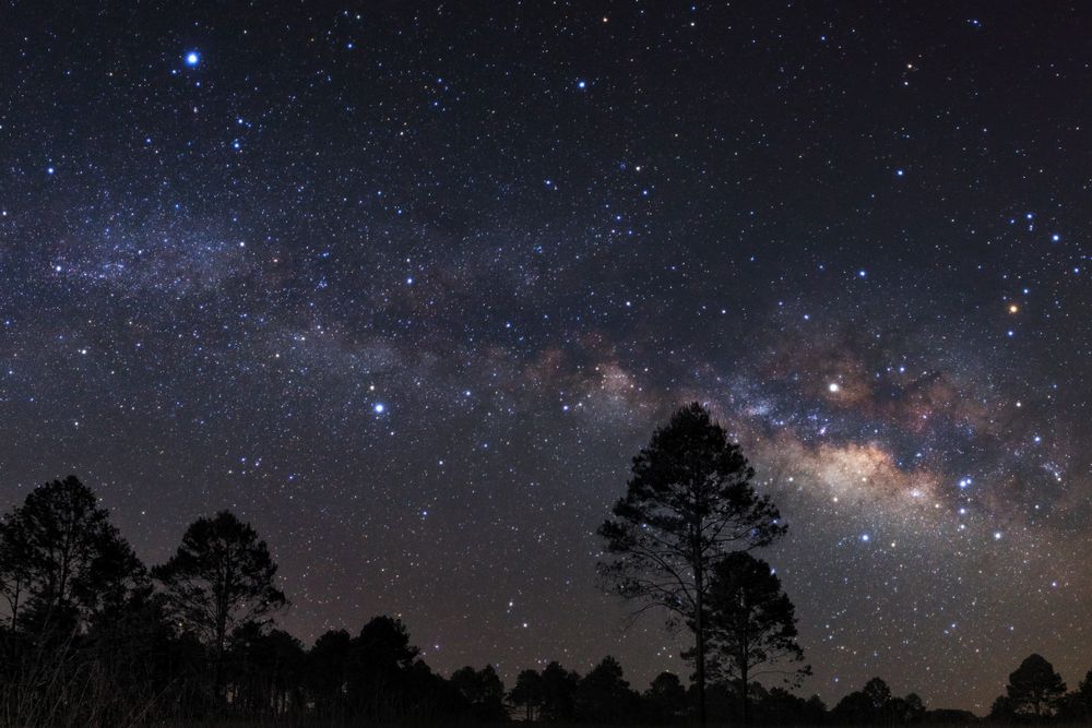 Night sky with stars and silhouette of a standing sporty man with raised up arms on high mountain.