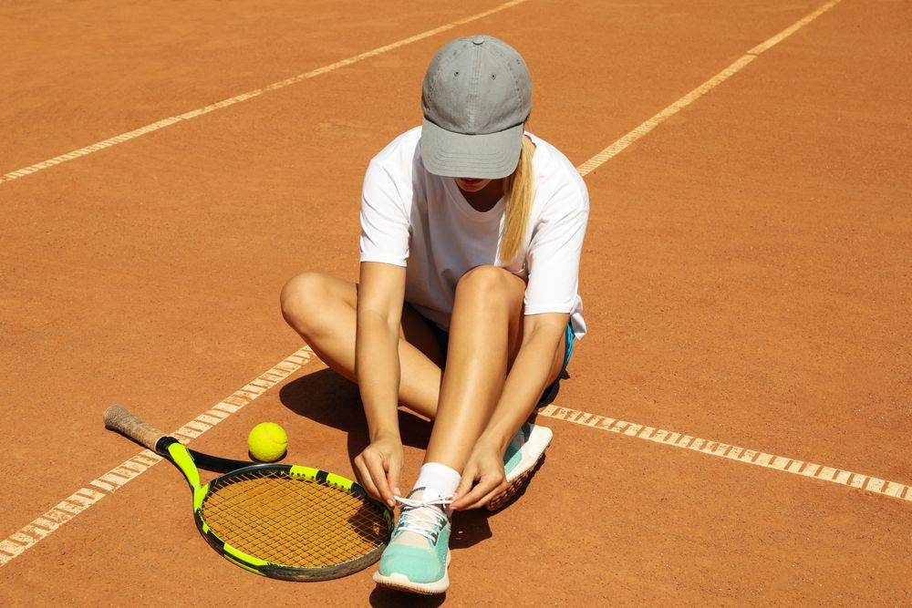 Woman ties shoelace on clay court with racket and and ball