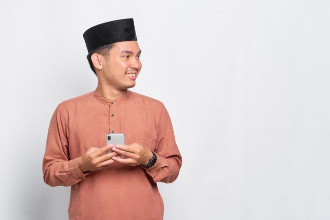 Muslim man in kufi hat holding mobile phone and looking to the side