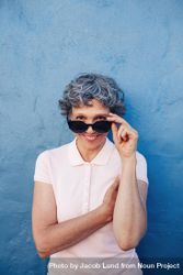 Portrait of attractive woman peeking over sunglasses against blue wall 0LNBP4
