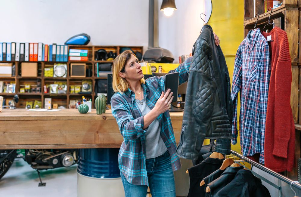 Blonde woman holding digital tablet while check leather jacket on second hand clothing store