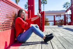 Side view of smiling young woman in red sitting taking selfie on mobile phone outdoors on a bright day