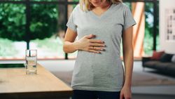 Young Adult Woman with Blond Hair Wearing Gray V-Neck T-Shirt, Rubbing Her Stomach After Food.