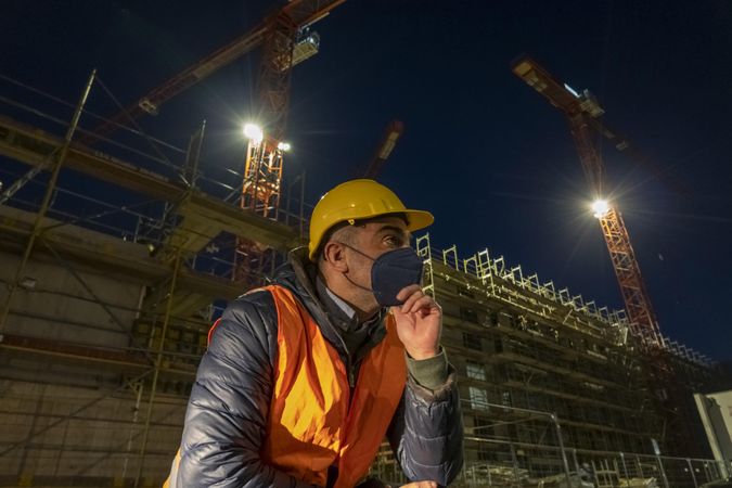 Construction worker with facemask sitting beside a building under construction at night