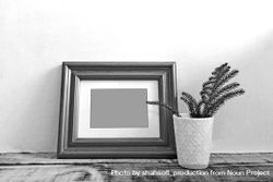 B&W shot of wooden picture frame leaning against wall next to plant in vase mockup 5R7WD4