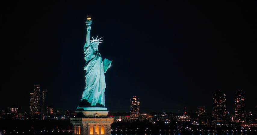 Panoramic Aerial Image of an American Symbol of Freedom. Cinematic Helicopter Pass by the Statue of Liberty at Night. Landmark New York Monument with Manhattan Skyscrapers in the Background