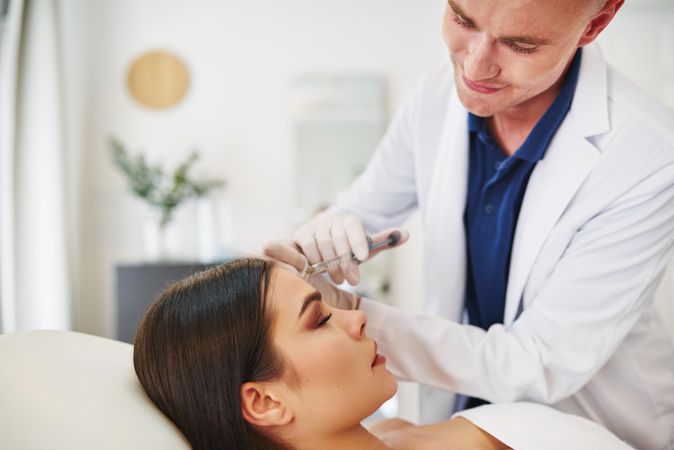 Side view of smiling male dermatologist injecting treatment into brunette patient’s face