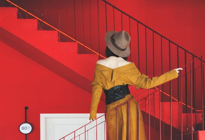 Back view of woman in yellow dress and gray hat standing on stairs in red room
