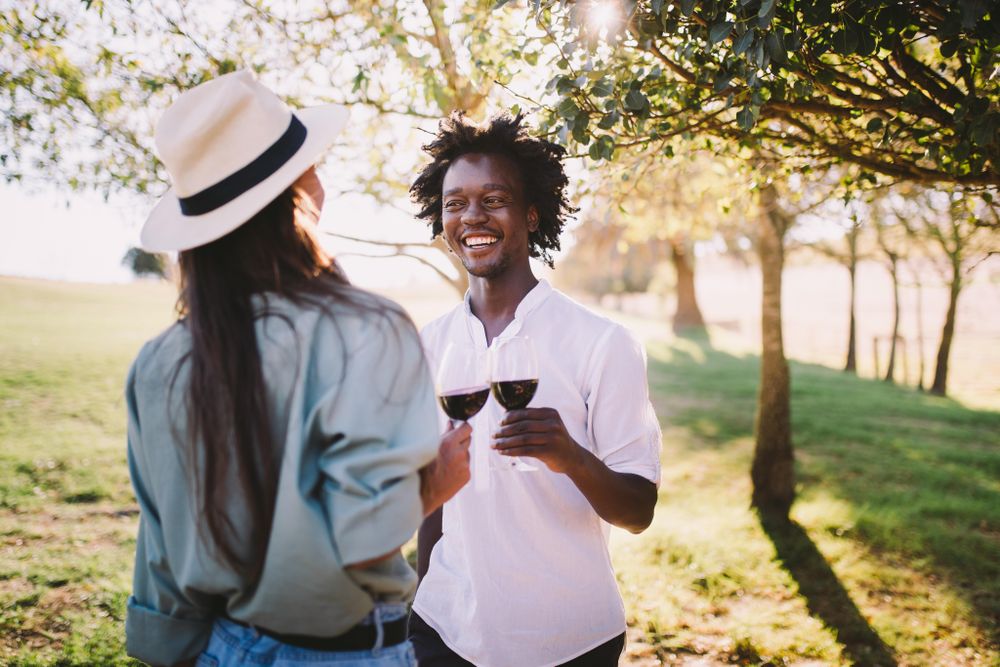 A man and woman holding red wine standing among trees on a sunny day