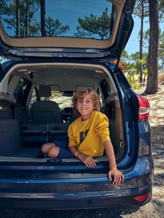 Smiling boy sitting in trunk of parked car in forest