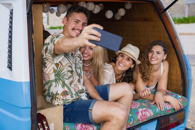 Smiling man taking selfie with girlfriends comfortable placing on trunk of blue car