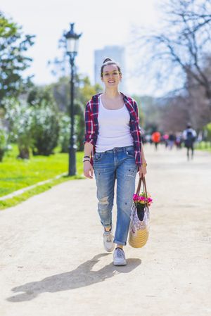 Front view of a young smiling hipster woman walking in a park in sunny day while holding a wicked basket