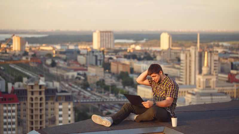 Man sitting on roof over looking skyline working on laptop with takeaway drink