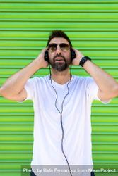 Man wearing sunglasses and holding headphone in front of green background beVlP0