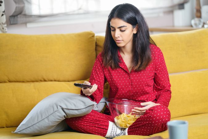 Female in red pajamas sitting on sofa with bowl of chips and remote control