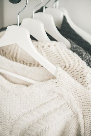 Close up of light to dark knitted sweaters on hangers, light background, vertical composition