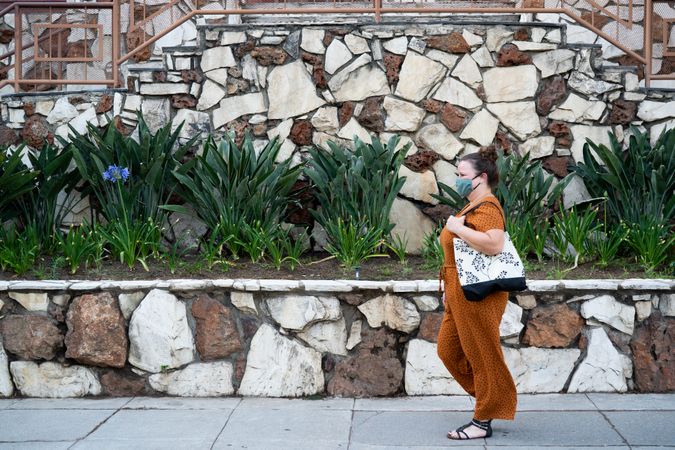 Side view of woman walking in front of stone wall carrying tote bag and wearing a mask