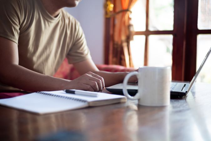 Asian Male with shirt working from home in living room with book, laptop and cup of coffee.