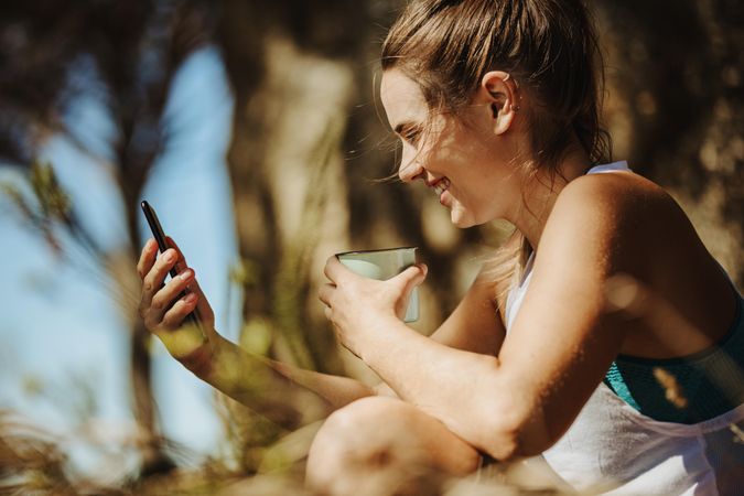 Smiling young woman looking at her phone and drinking coffee outdoors while on a hike