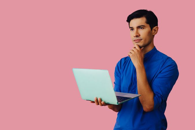 Contemplative Latino man holding laptop in pink room, copy space