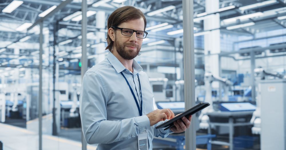 Portrait of a Handsome Adult Engineer Wearing Glasses, Using Tablet Computer and Looking at Camera. Technician in a Shirt Working in a Modern Technology Factory with Robotis Assembly Arms