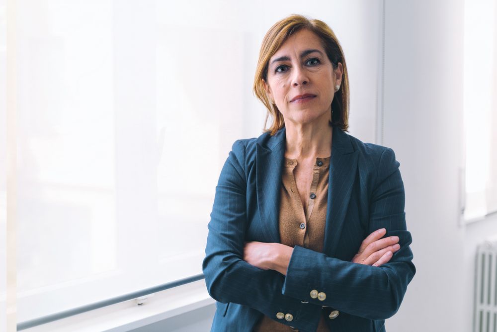 Businesswoman in her 50s, dressed in casual office attire, standing near window with arms crossed, looking seriously at camera