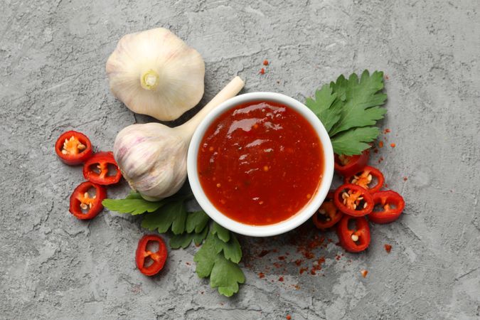 Bowl with chilli sauce, pepper and garlic on gray background