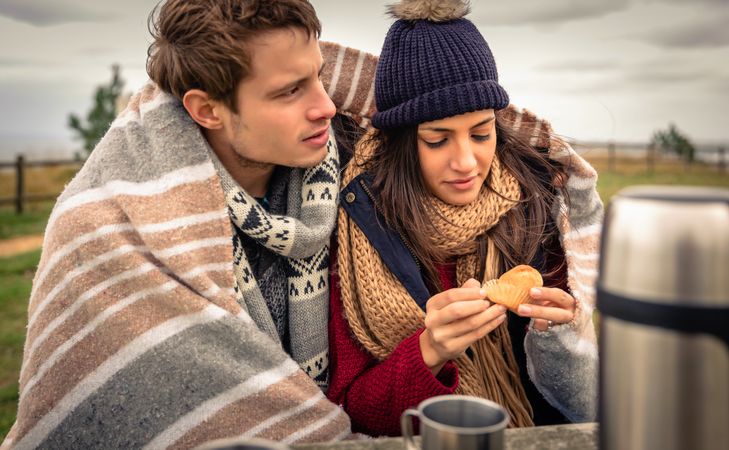Cute couple wrapped in blanket on cold park bench with coffee flask and muffin