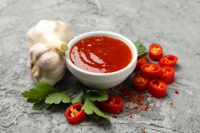 Bowl with chilli sauce, pepper and garlic on gray background