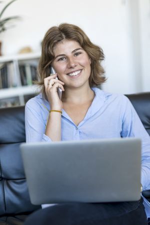 Smiling woman sitting on sofa at home using a laptop and chatting on phone