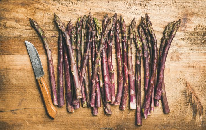 Scattered pieces of washed purple asparagus, on wooden board with knife