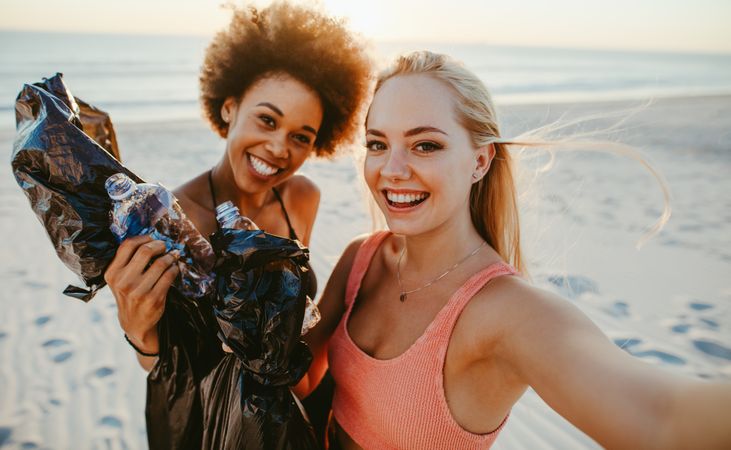 Two young women holding garbage bags and taking selfie at the beach area