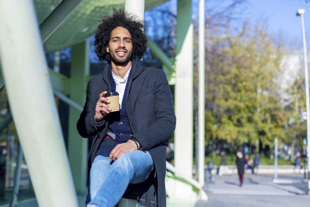 Front view of cheerful Black man in jacket with warm beverage balancing between poles outside