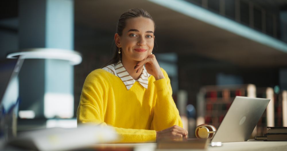 White Female Student Working on Her School Thesis on a Laptop Computer in a Modern Library. Young Smart Woman Looking at Camera, Smiling.