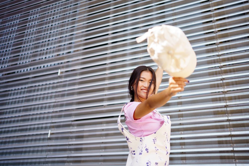 Smiling Chinese woman in floral coveralls throwing her handbag in front of metal wall
