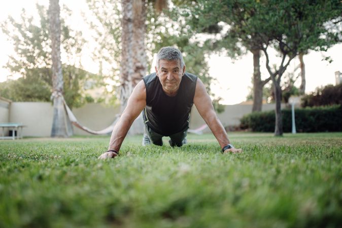 Cheerful older man pushing ups on green grass in park