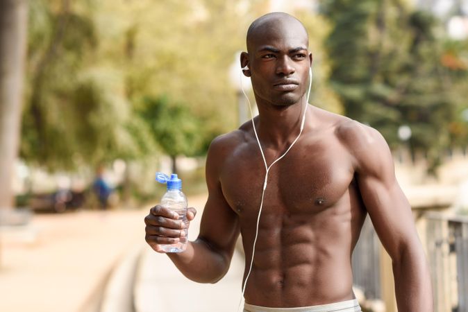 Healthy male with shirt off drinking from a water bottle in an outdoor park